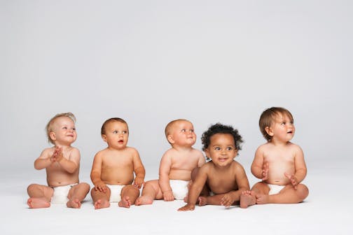 Row of multiethnic babies sitting side by side looking away isolated on gray background.