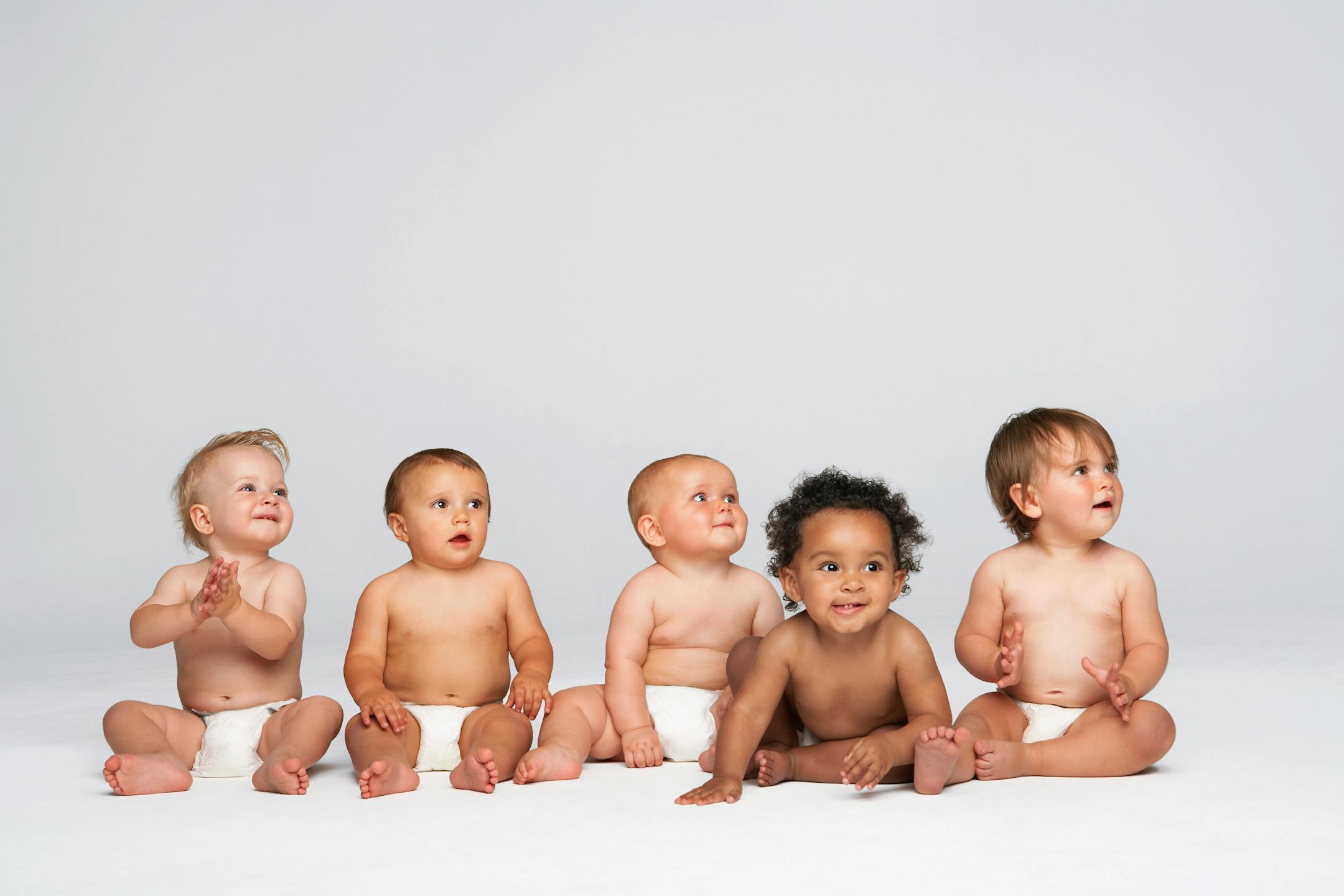 Row of multiethnic babies sitting side by side looking away isolated on gray background.