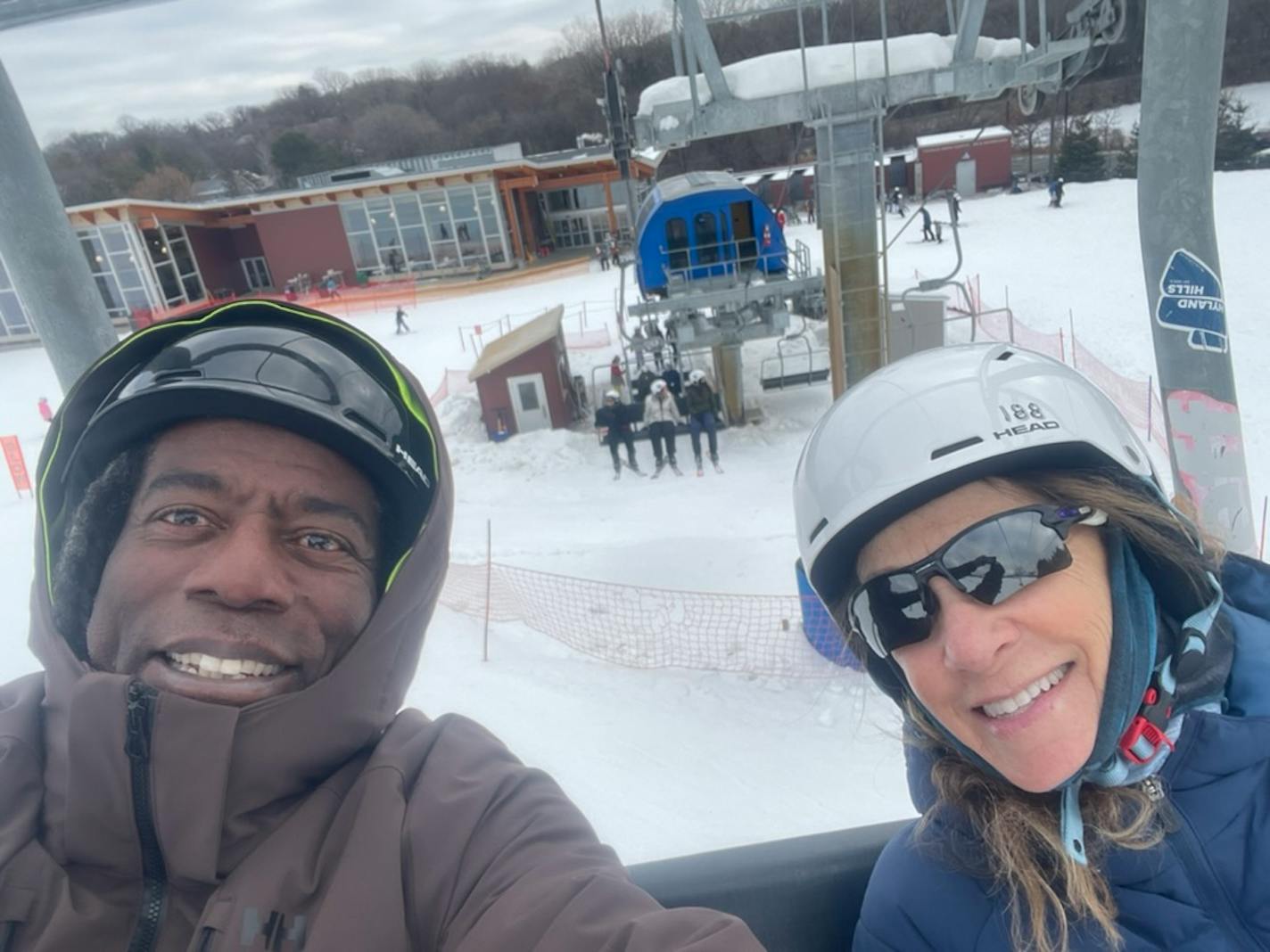 A man and a woman smile for a selfie taken on a ski lift. A modern chalet building and snow are seen in the backround.