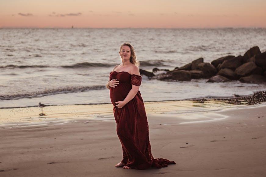 Renee Nicole Good stands on a beach at sunset. She wears a red gown and is smiling into the camera.