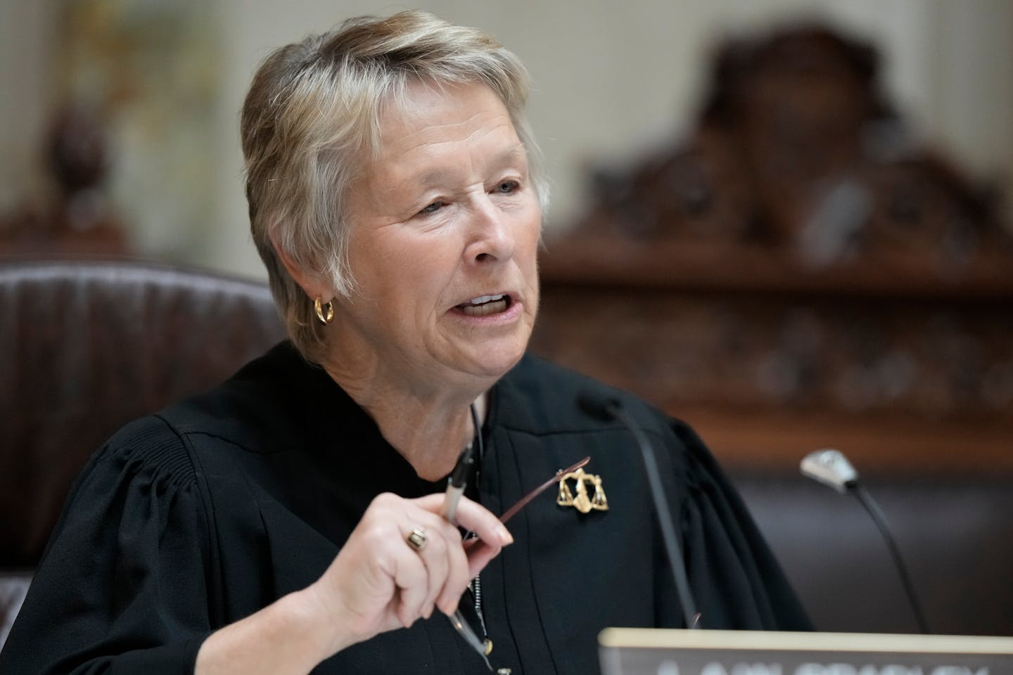 Wisconsin Supreme Court Justice Ann Walsh Bradley at bench wearing black judicial robe with scales of justice pin and holding glasses in right hand.