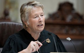 Wisconsin Supreme Court Justice Ann Walsh Bradley at bench wearing black judicial robe with scales of justice pin and holding glasses in right hand.