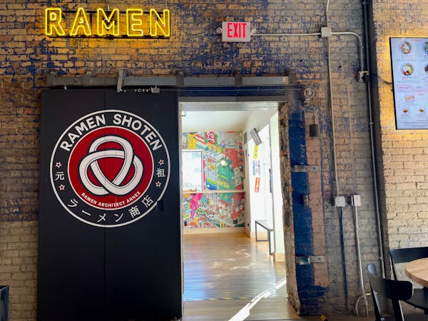 A brick wall and the sliding metal door adorned with Ramen Shoten. There's a yellow neon sign that reads "Ramen" above the door.