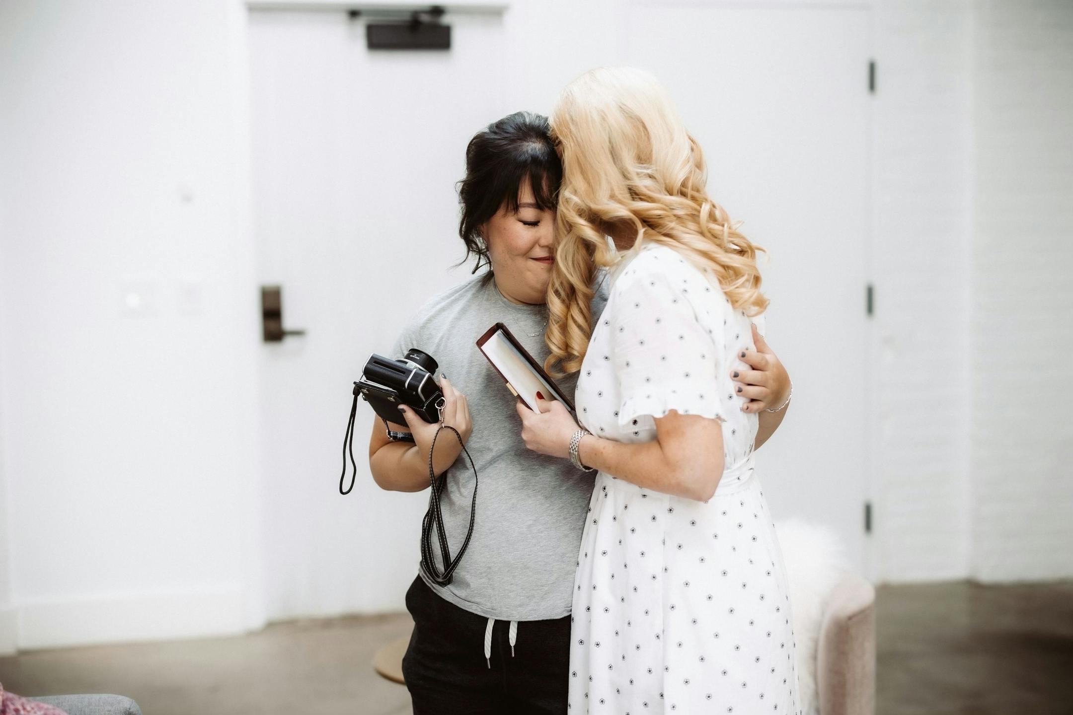 A woman in her 30s in sweat pants and an elegant updo closes her eyes as she hugs a blond woman wearing a casual white dress.