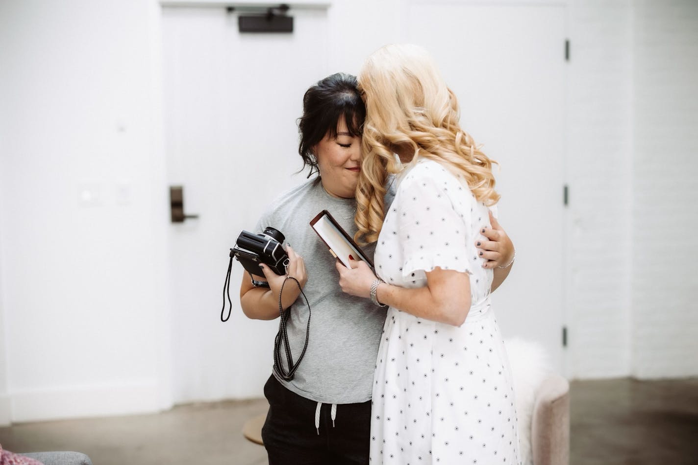 A woman in her 30s in sweat pants and an elegant updo closes her eyes as she hugs a blond woman wearing a casual white dress.