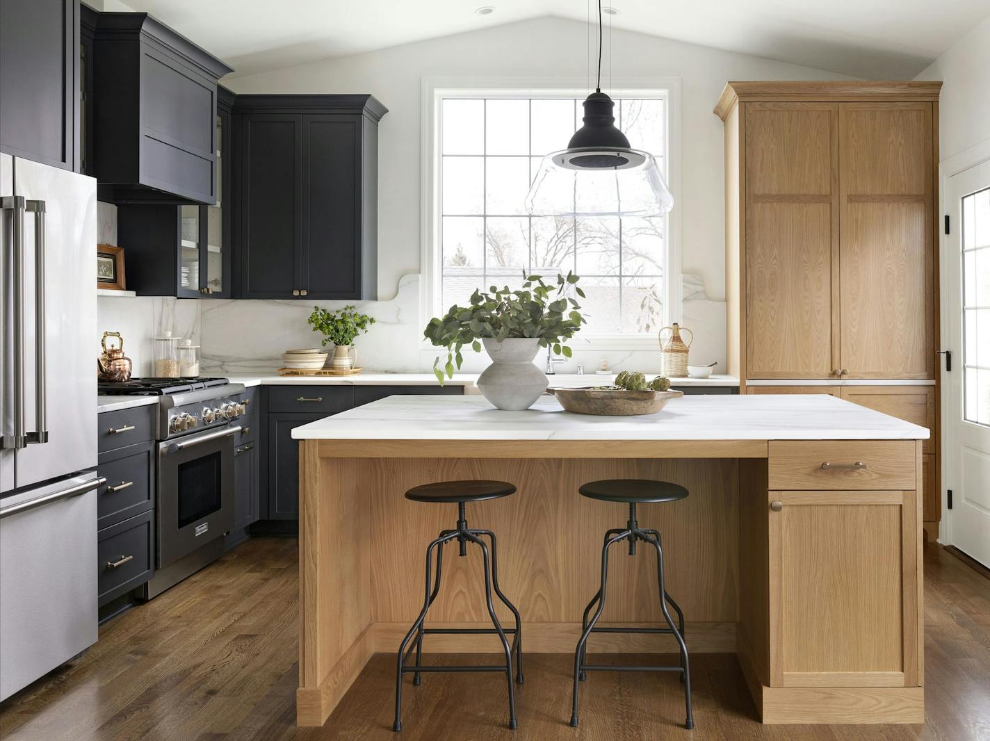 Cabinetry is a combination of white oak with a light natural finish and painted dark charcoal; some cabinets have glass doors for display. A built-in cabinet next to the sink provides pantry-like storage for small appliances, in addition to food staples.