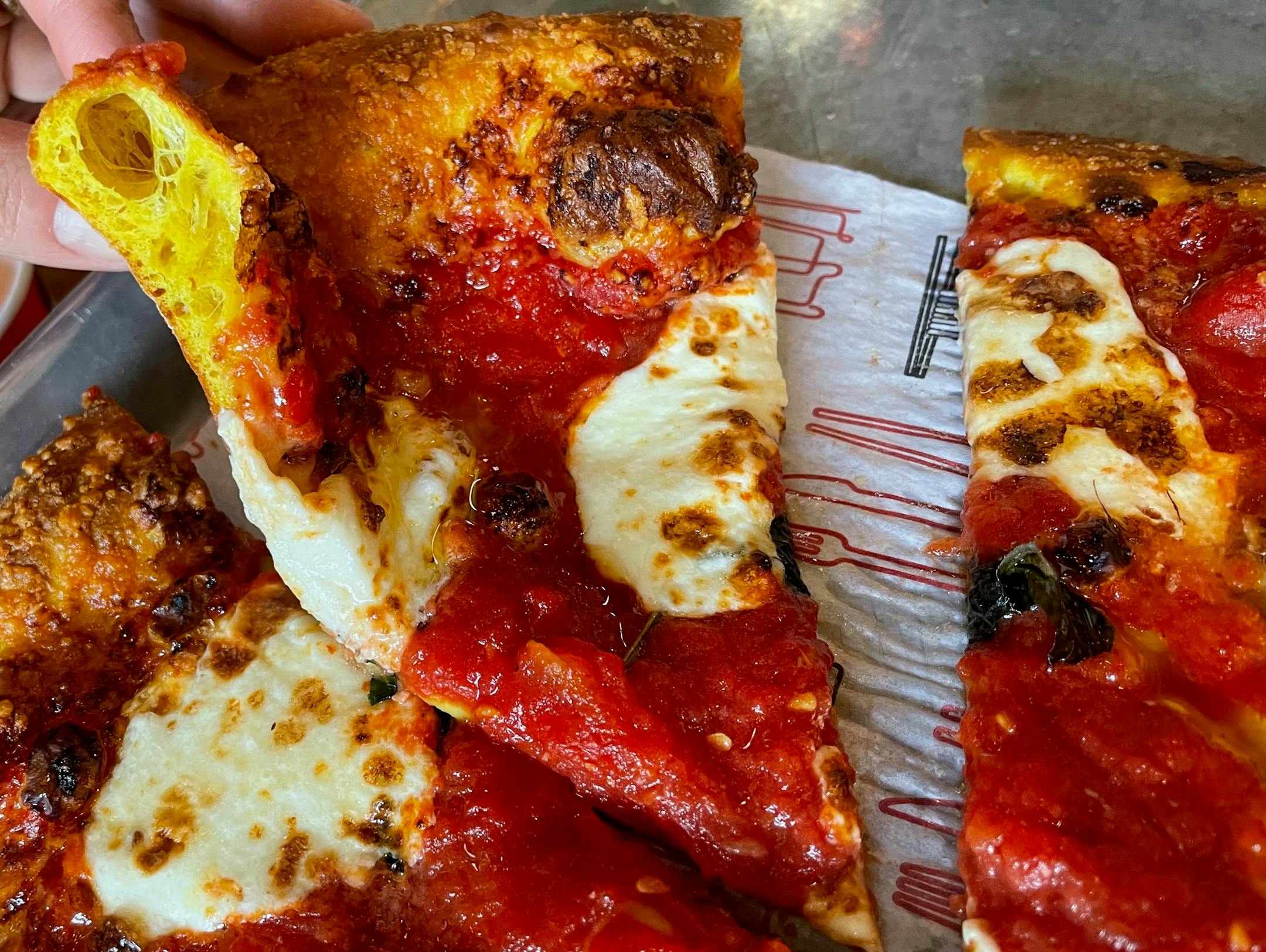 A hand picks up a slice of pizza with crushed tomatoes, basil and blobs of mozzerella cheese. The crust has a distinctive yellow color on the inside.