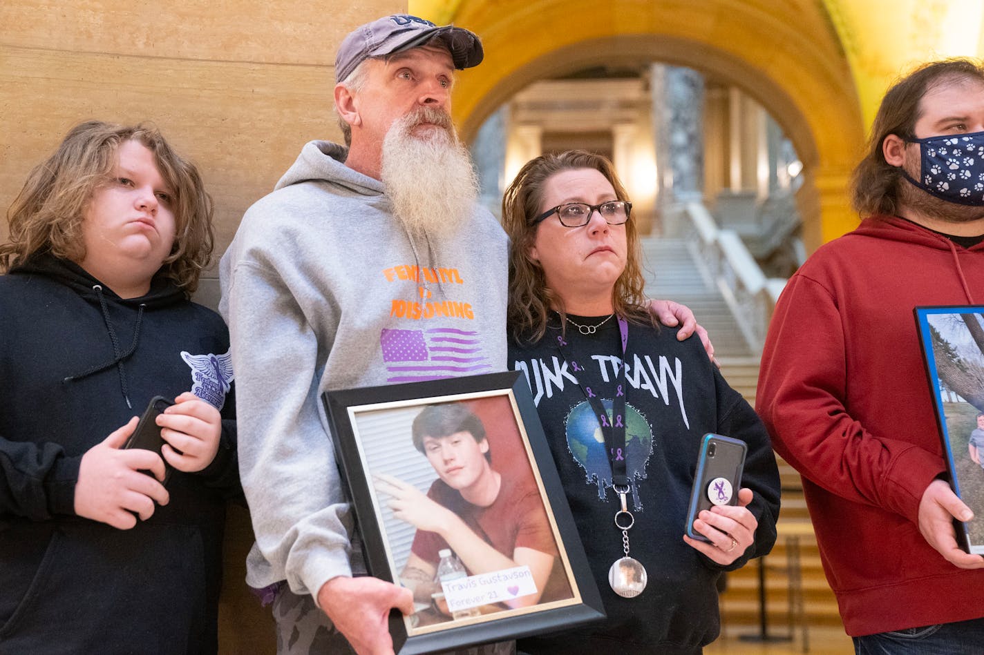 Jason Lange holds a photo of his stepson, Travis Gustavson, while putting his arm around his wife and Travis' mother, Kim Gustavson, at an event calling for increased legislative action regarding fentanyl Tuesday, Feb. 21, 2023 at the Minnesota State Capitol in St. Paul. ]