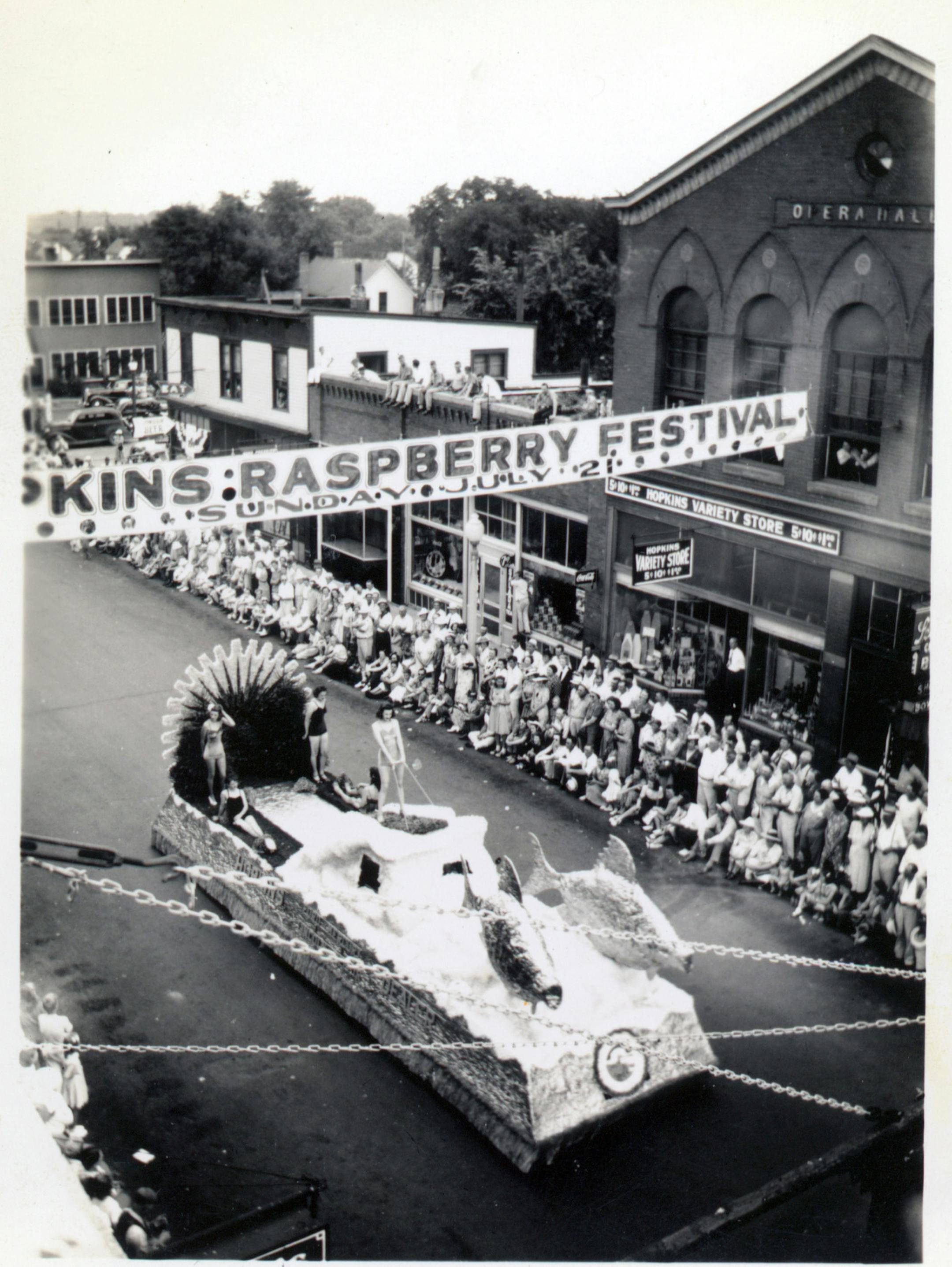 The caption (provided by the Hopkins Historical Society) is as follows: B/W photograph of 1946 Raspberry Festival parade taken from building on south side of Excelsior Avenue. Opera Hall, festival banner, float with bathing beauties (one skiing using two fish). Spectators on the street and atop buildings on north side. ORG XMIT: MIN2013071516233360