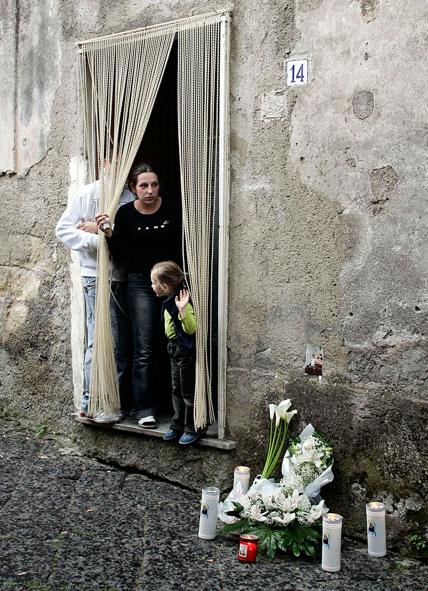 Flowers and candles were left near the home where 6-year-old Karolina, whose photo was taped on the wall at right, was killed by a gunshot when an Italian man fired through the door, in San Paolo Belsito, near Naples, Italy, Saturday, May 5, 2007. The man, who later turned himself in, had previously argued with two Polish immigrants and later returned to fire two gunshots in the home of one of them, killing the girl. The woman at the door is the mother of Karolina. (AP Photo/Salvatore Laporta) O