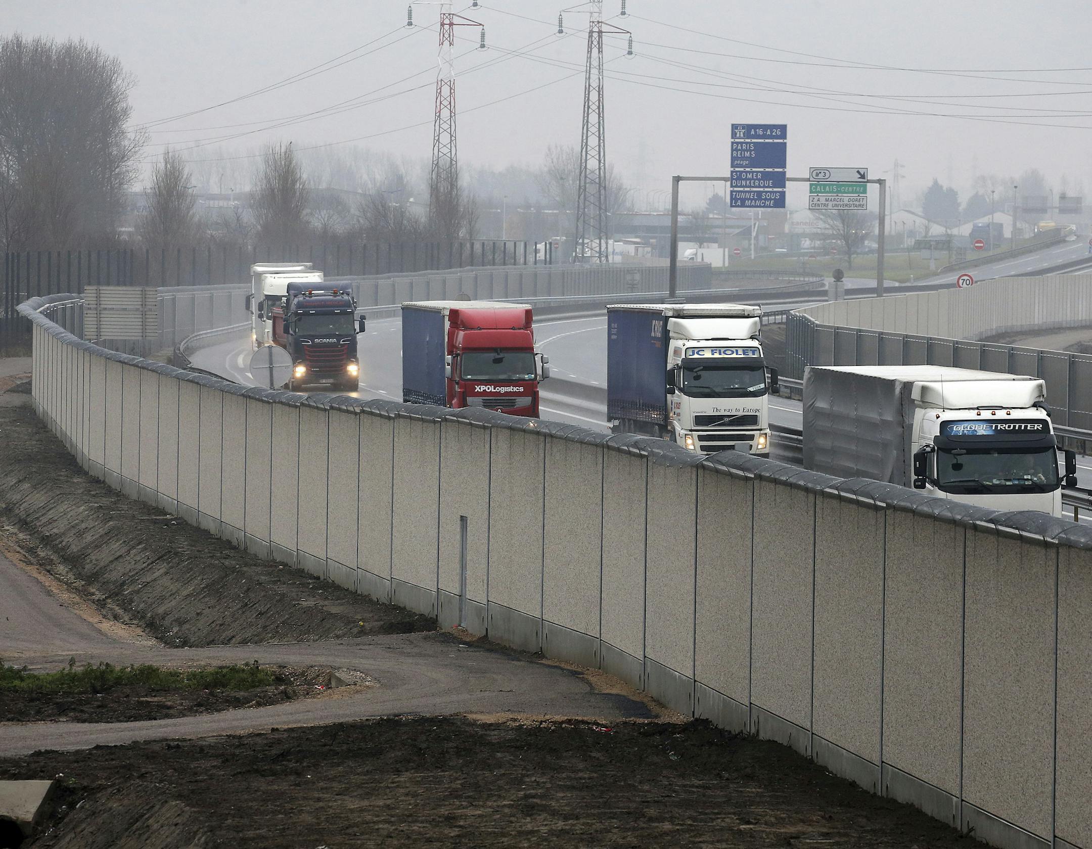 File - In this Tuesday, Dec. 13, 2016 file photo, trucks drive past a 4 meters (13-foot) high wall along the road leading to the Calais port, outside Calais, northern France. A concrete wall, one kilometer (over half a mile) long and four meters (13-feet) high, is the finishing touch on an already elaborate defense system against migrants in the northern French port city of Calais. (AP Photo, File)
