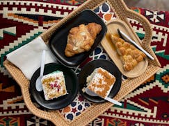 A wicker tray holds two pieces of tres leches cake, an almond croissant and a honeycomb bread slice. The tray is placed on a red woven banquette.