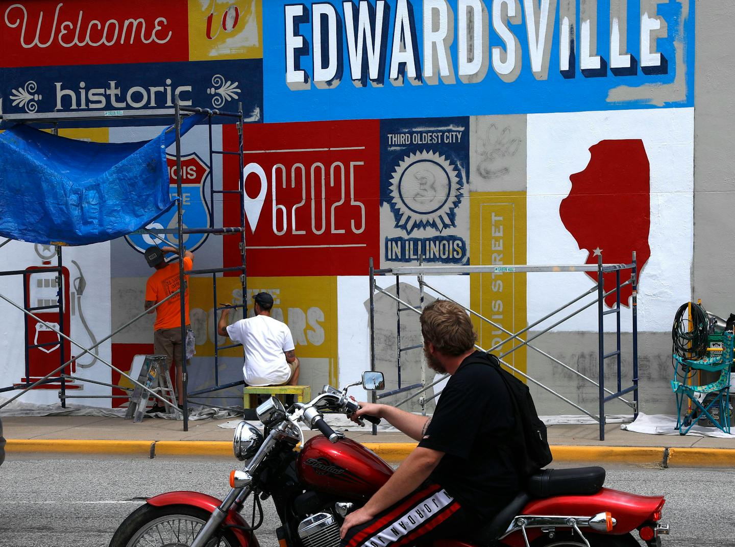 A motorist watches owner Daniel Ricketts, left, and muralist Jerome Lamke with St. Louis Sign &amp; Mural, hand-paint a Route 66 mural on a building at the southeast corner of East Vandalia and South Main streets on Monday, July 25, 2022, in Edwardsville, Illinois. The mural is one of 11 murals highlighting Route 66 in southern Illinois towns. (Laurie Skrivan/St. Louis Post-Dispatch/TNS) ORG XMIT: 55889262W