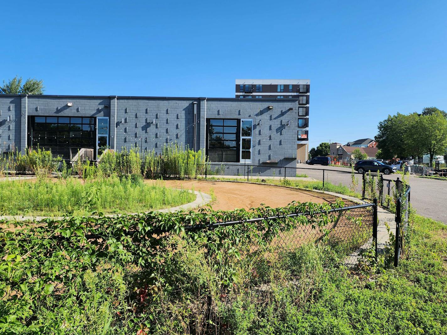 A gray brick building with a weedy and overgrown dirt bike track in front.