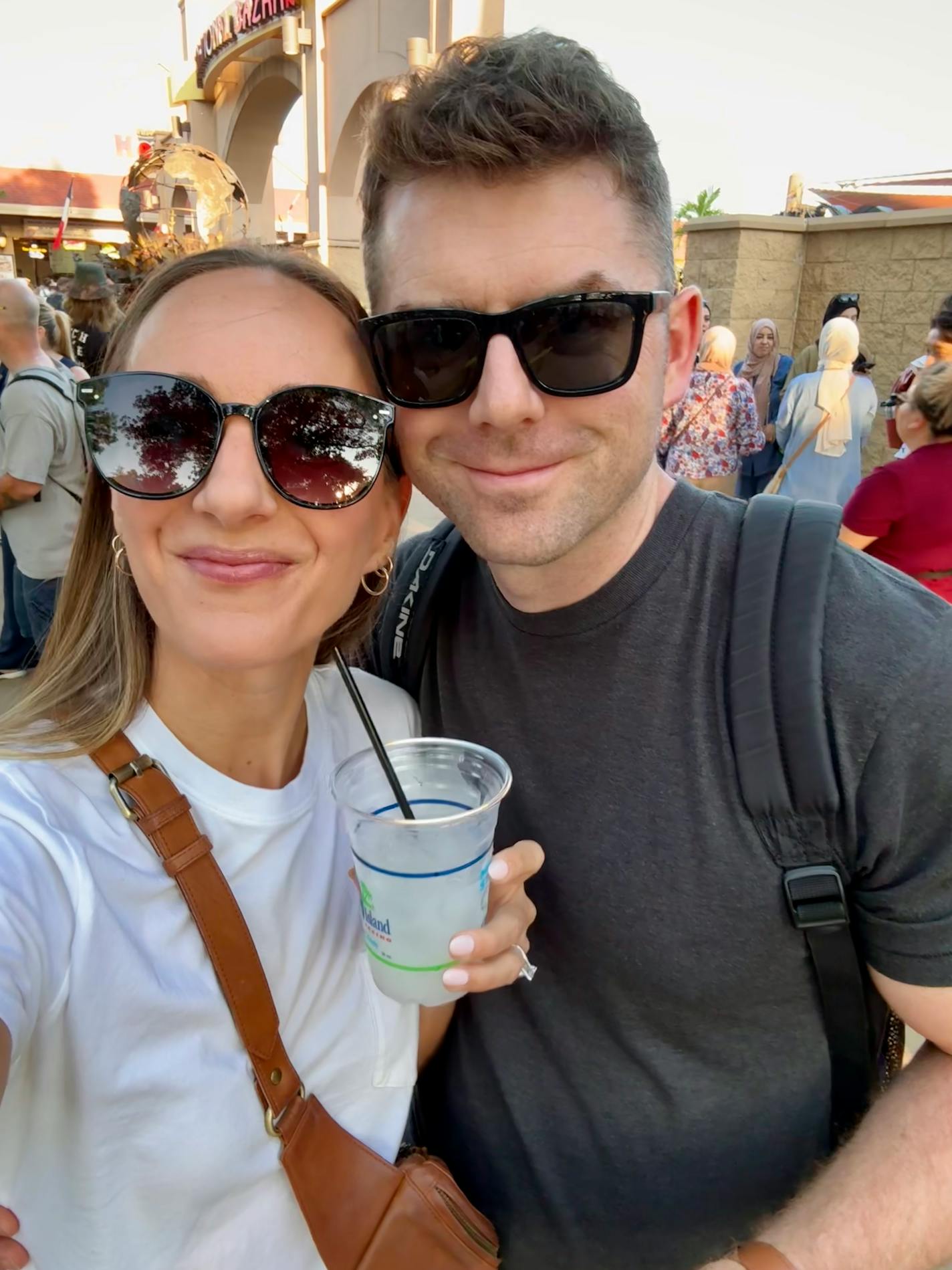 A couple wearing sunglasses pose for a selfie. Woman is holding a clear plastic cup with straw and a clear beverage.