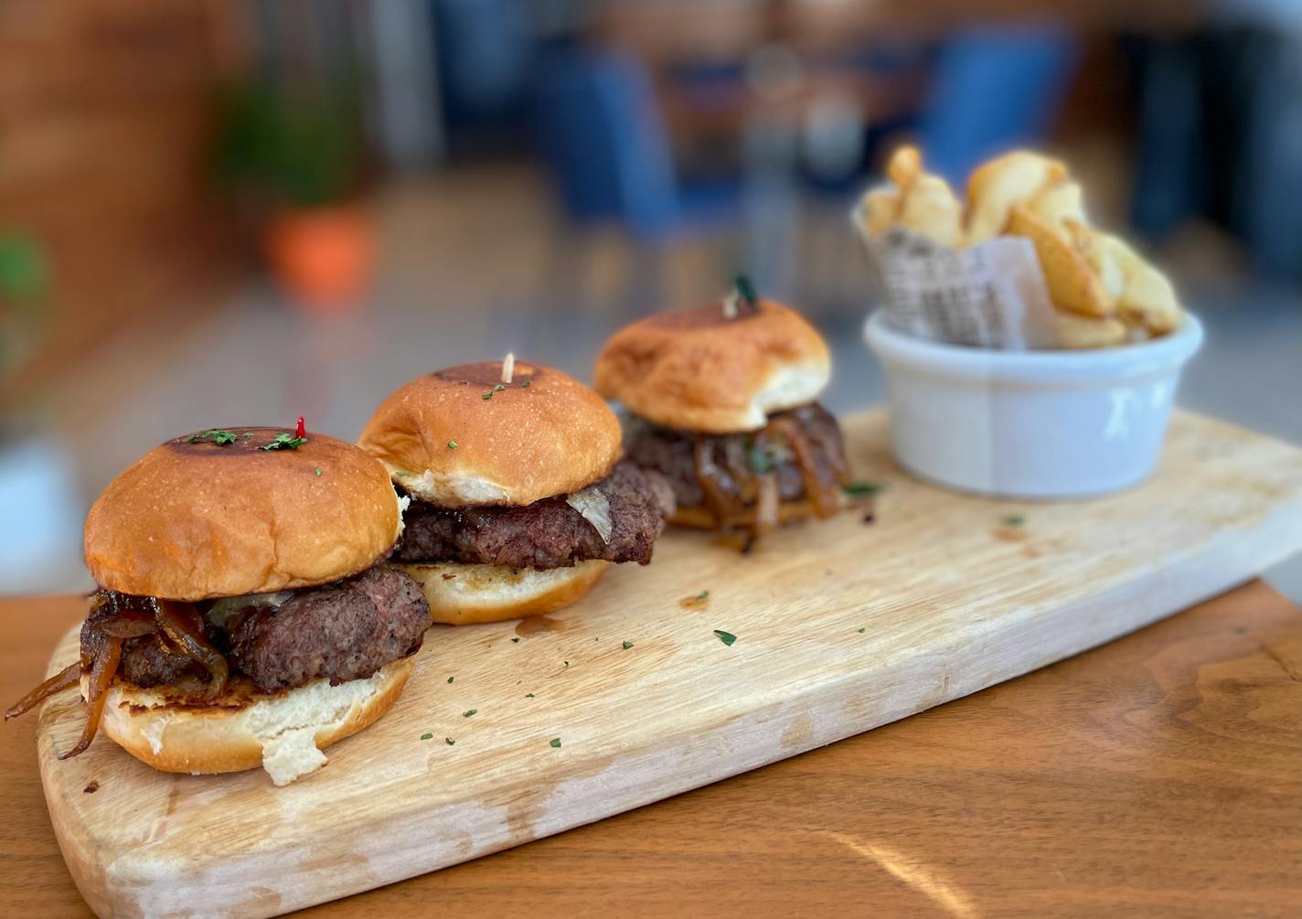 Three small burgers topped with caramelized onions are lined up on a wooden board with a cup of French fries in the background.