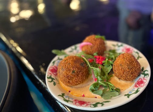 A white with floral design plate holds three large, fried boudin balls with a centerpiece of arugula, in a pool of pimento-colored sauce.