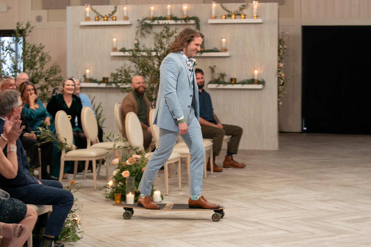 A man in a powder blue suit skates his longboard past wedding guests with a big smile on his face.