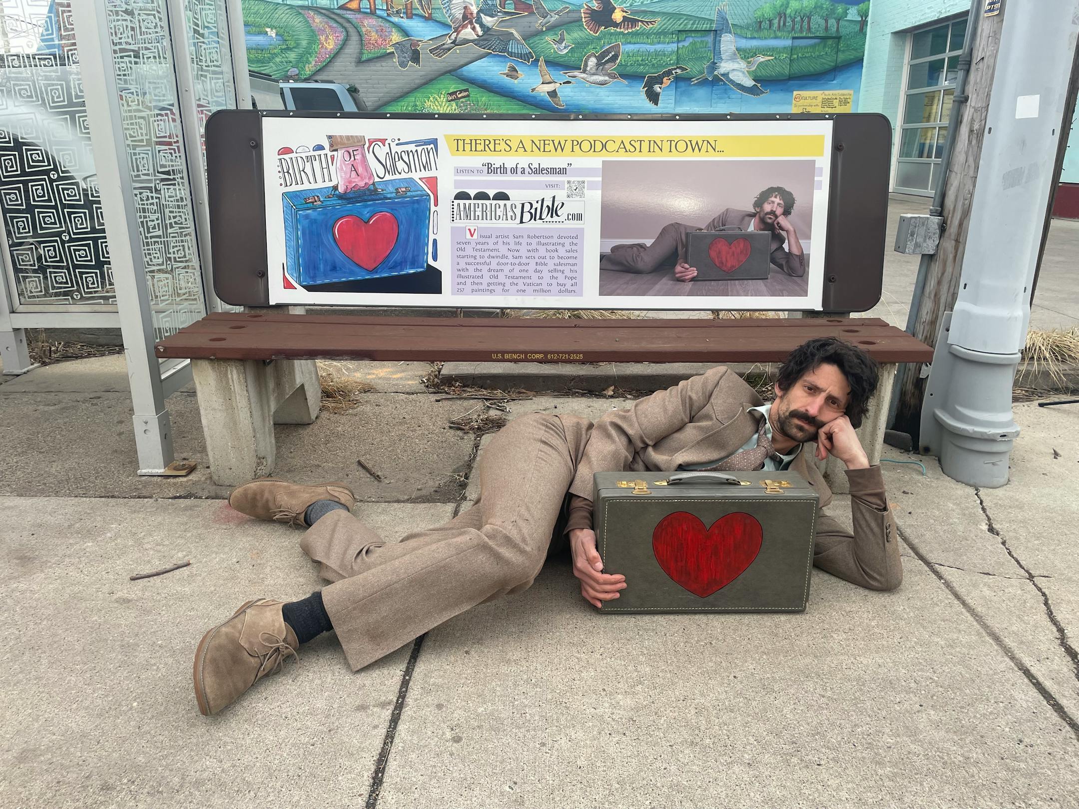 Artist Sam Robertson in front of a bus-bench advertisement about his illustrated Old Testament, a seven-year artistic project that is now the subject of a narrative podcast.