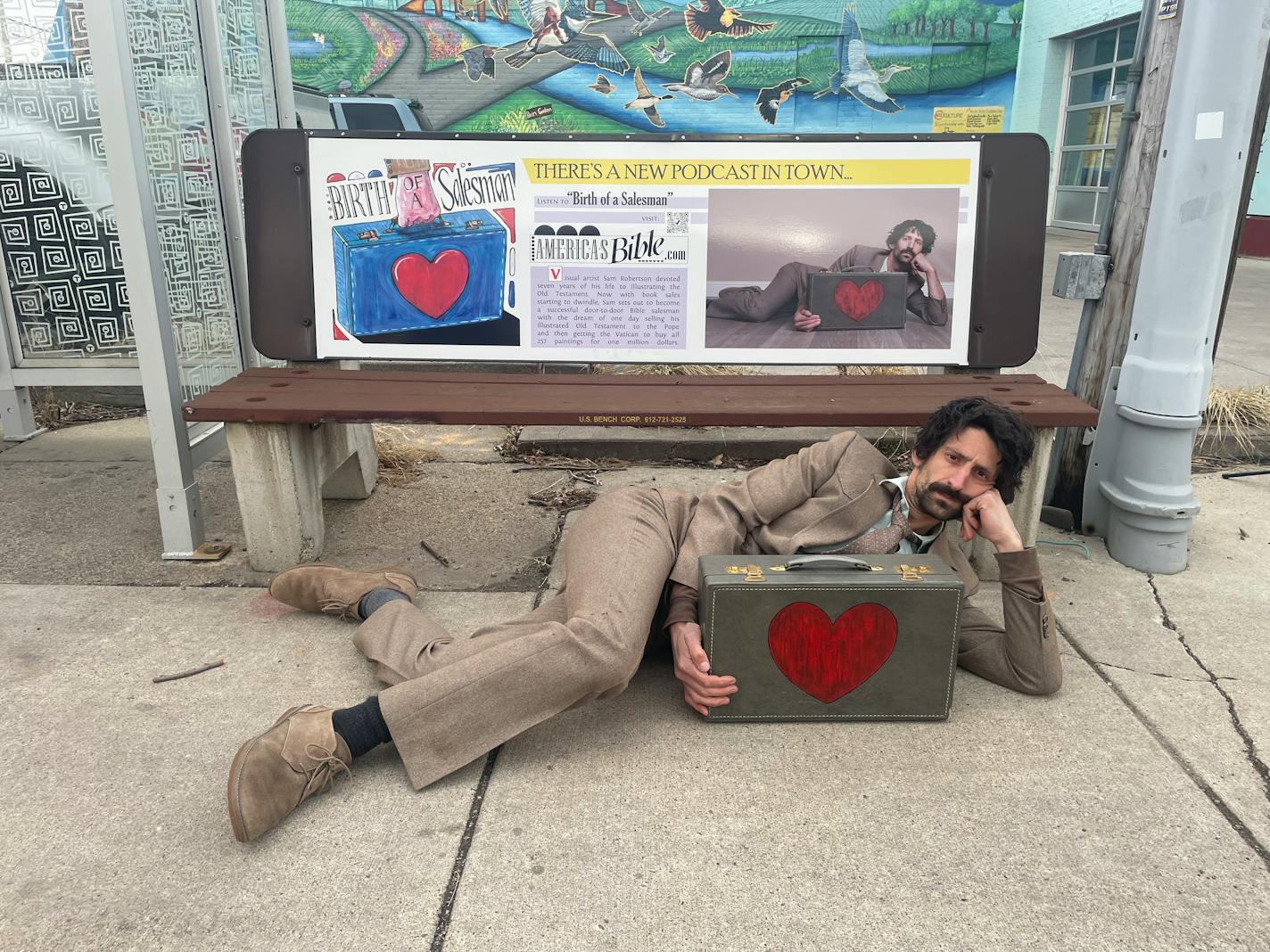 Artist Sam Robertson in front of a bus-bench advertisement about his illustrated Old Testament, a seven-year artistic project that is now the subject of a narrative podcast.