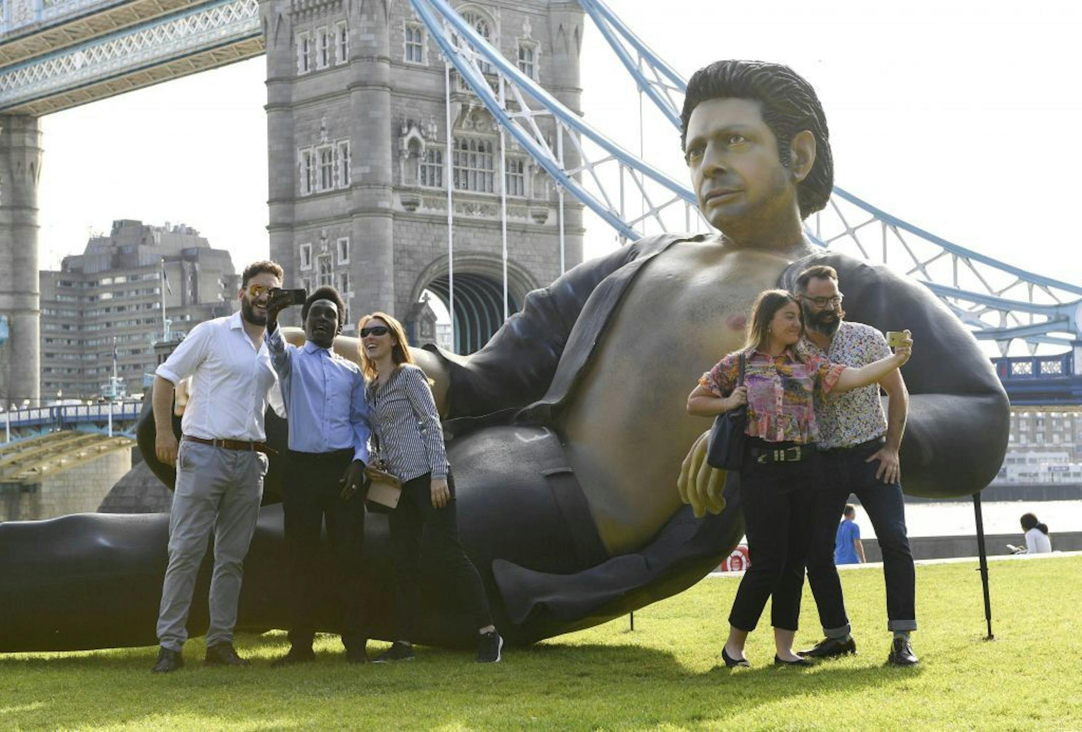 People take photos by a 25ft statue of actor Jeff Goldblum in a pose from a scene in the first Jurassic Park movie, which has been created by a TV channel to celebrate the film's 25th birthday, at Potters Fields Park, London, Wednesday July 18, 2018. Tower Bridge in the background.