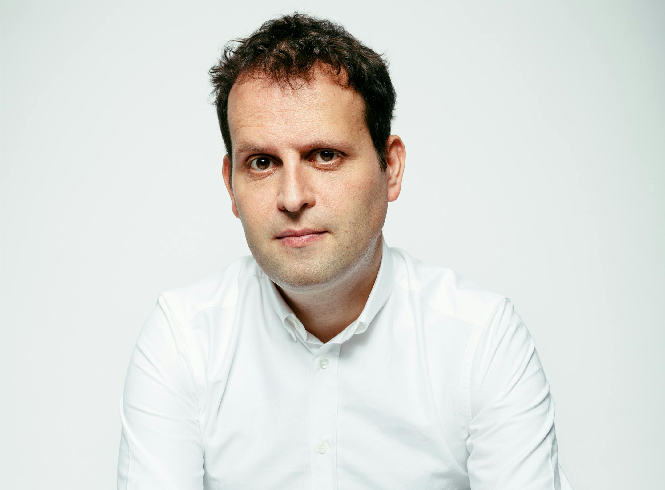 against a white background, photo of author Adam Kay in a white button-down shirt