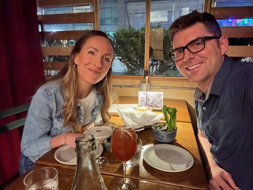 A young couple smile for a selfie at a restaurant, pictured next two two beverages.