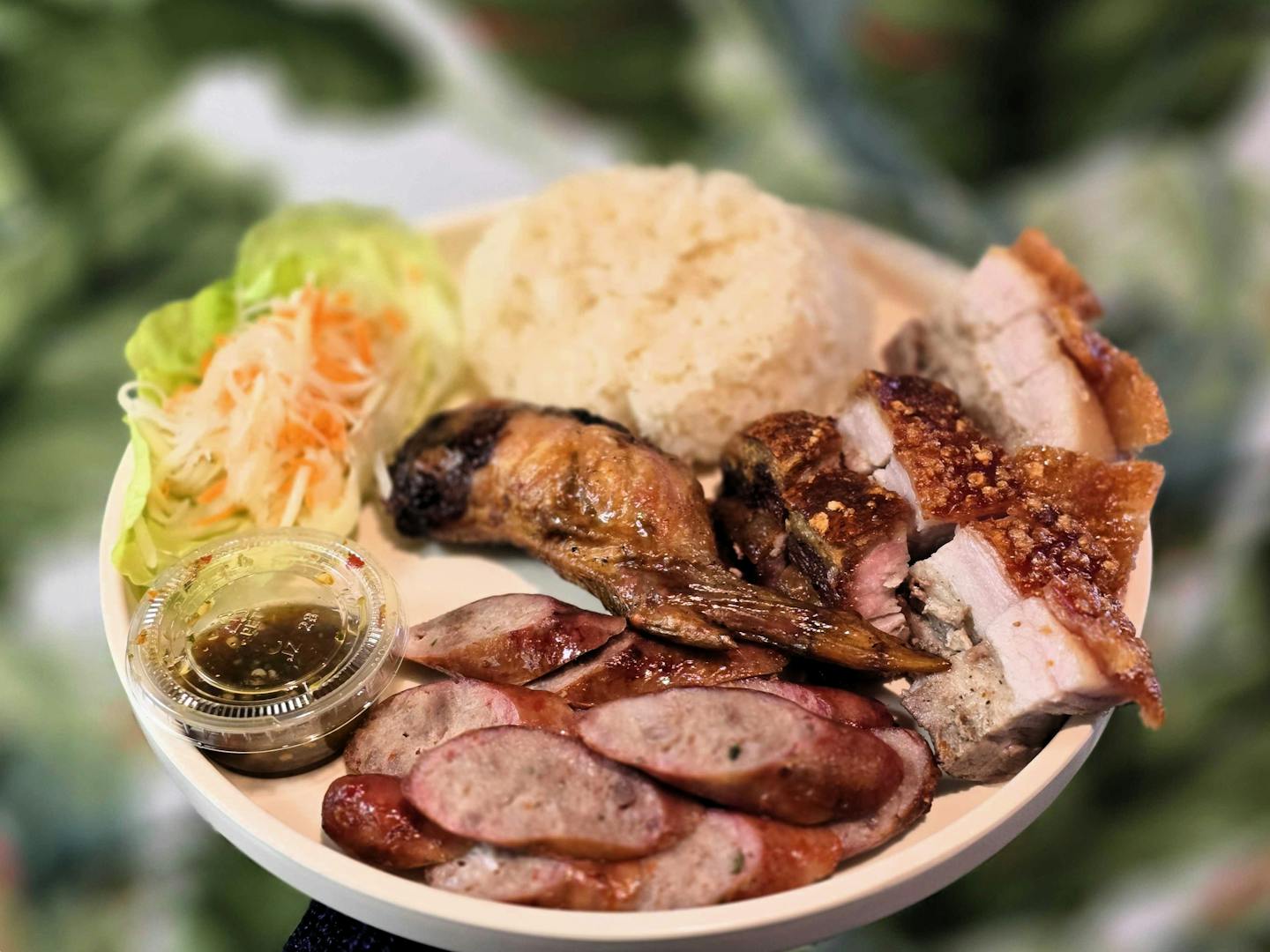 A plate with slices of Hmong sausage, a stuffed chicken wing and crispy pork belly, a mound of white sticky rice and shreds of white and orange papaya salad in a lettuce leaf