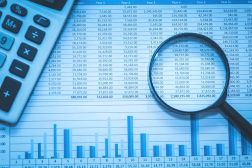 A stock image of a magnifying glass over a spreadsheet of dollar amounts