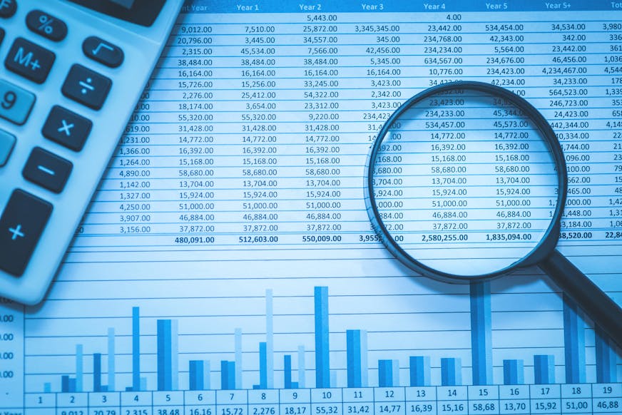 A stock image of a magnifying glass over a spreadsheet of dollar amounts