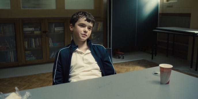 A boy in a track suit jacket half-smiles while sitting in a large table.