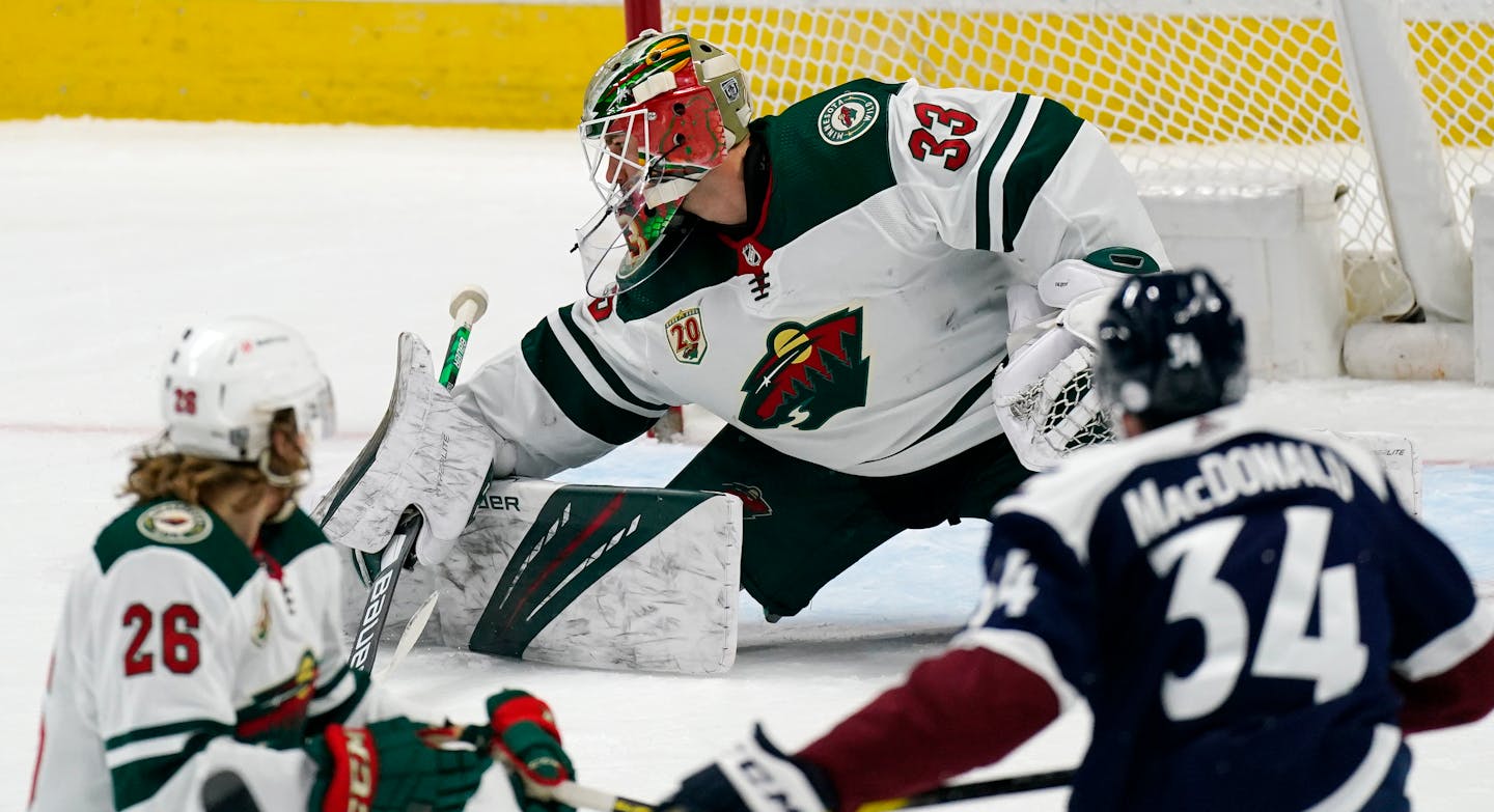 Minnesota Wild goaltender Cam Talbot, back, makes a stick save of a shot as right wing Gerald Mayhew, front left, and Colorado Avalanche defenseman Jacob MacDonald watch during the first period of an NHL hockey game Tuesday, Feb. 2, 2021, in Denver. (AP Photo/David Zalubowski)