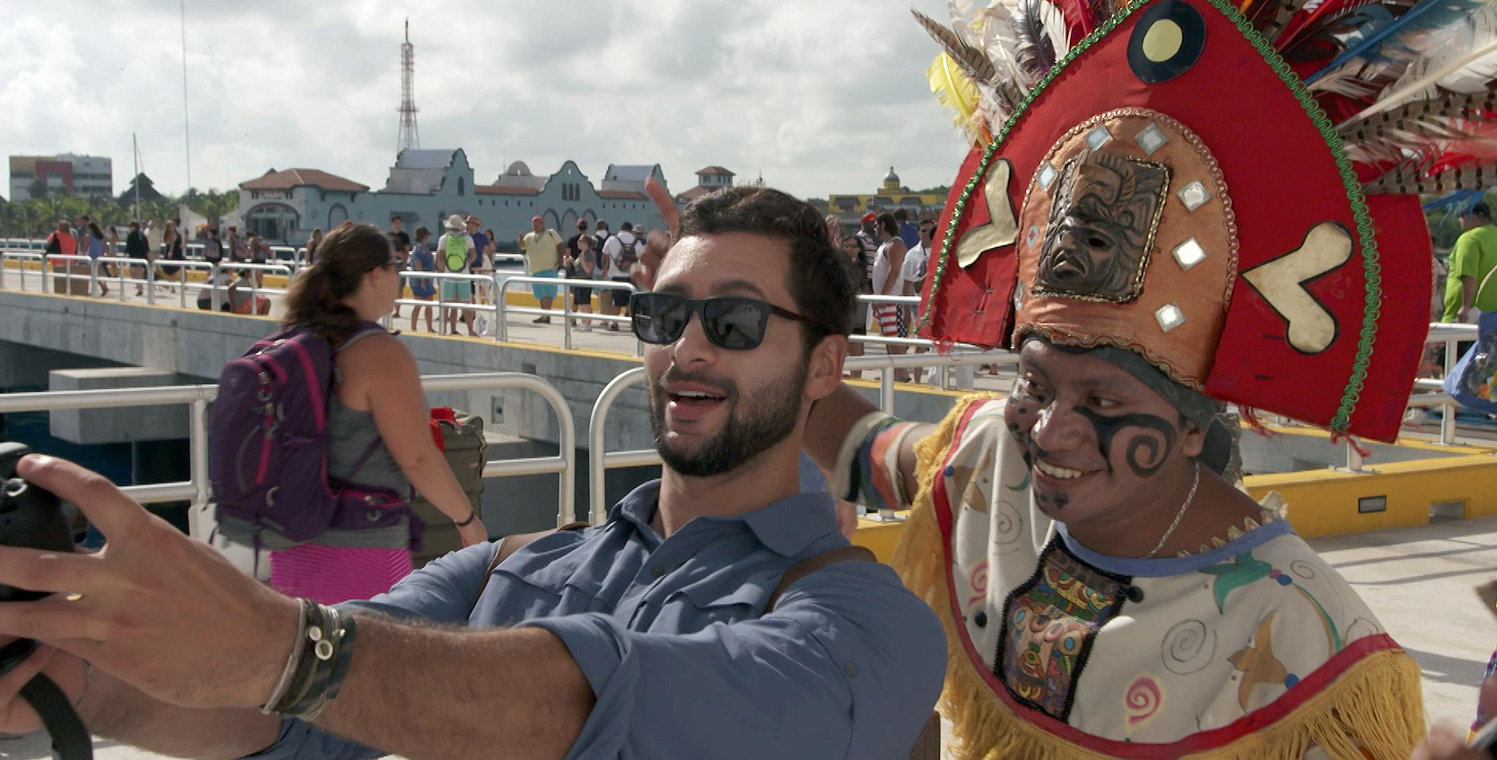 This undated photo provided by Carnival Corporation shows Josh Garcia in Playa del Carmen, Mexico, taking a selfie with a local resident dressed in costume to greet cruise passengers in a scene from a new show airing on NBC this fall called "The Voyager with Josh Garcia." The show is one of three new shows produced by Carnival Corp., showcasing vacation and travel connected to cruising. Josh Garcia hosts the show, which explores the history and culture of various ports through meetings with loca