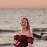 Renee Nicole Good stands on a beach at sunset. She wears a red gown and is smiling into the camera.