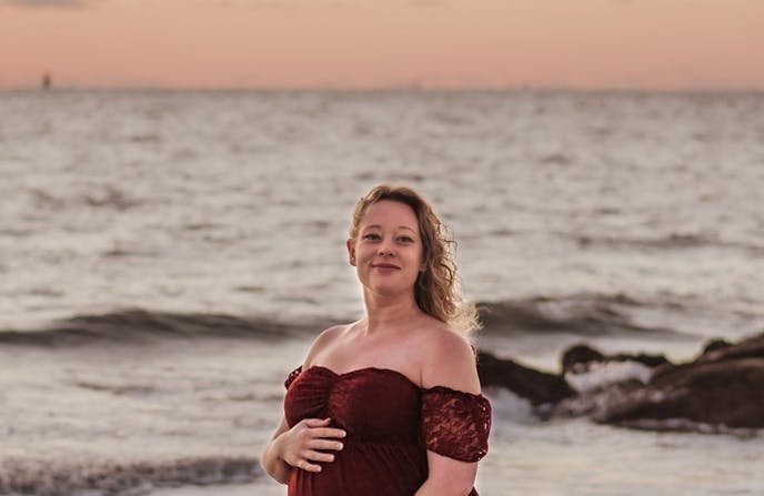 Renee Nicole Good stands on a beach at sunset. She wears a red gown and is smiling into the camera.