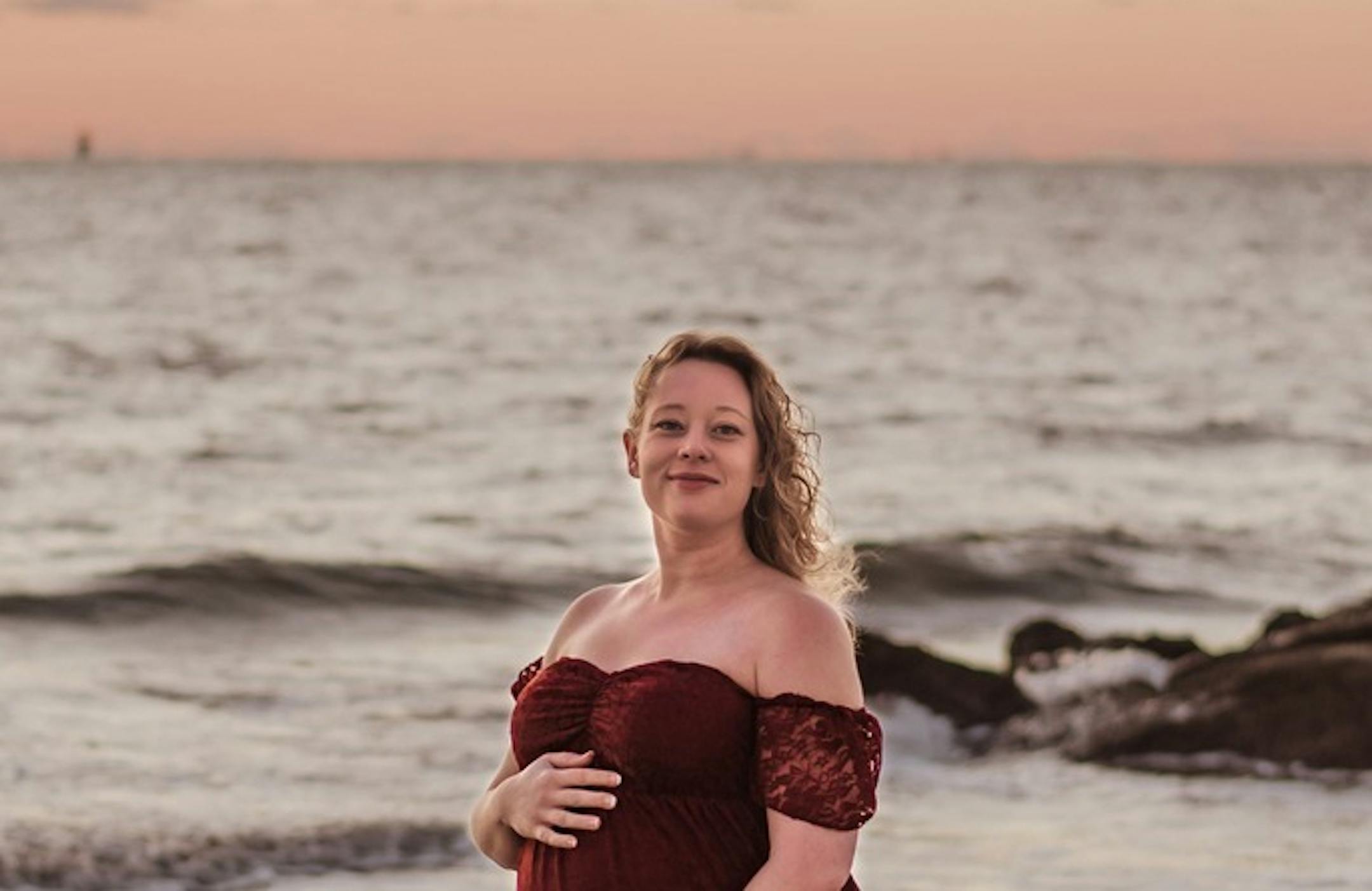 Renee Nicole Good stands on a beach at sunset. She wears a red gown and is smiling into the camera.