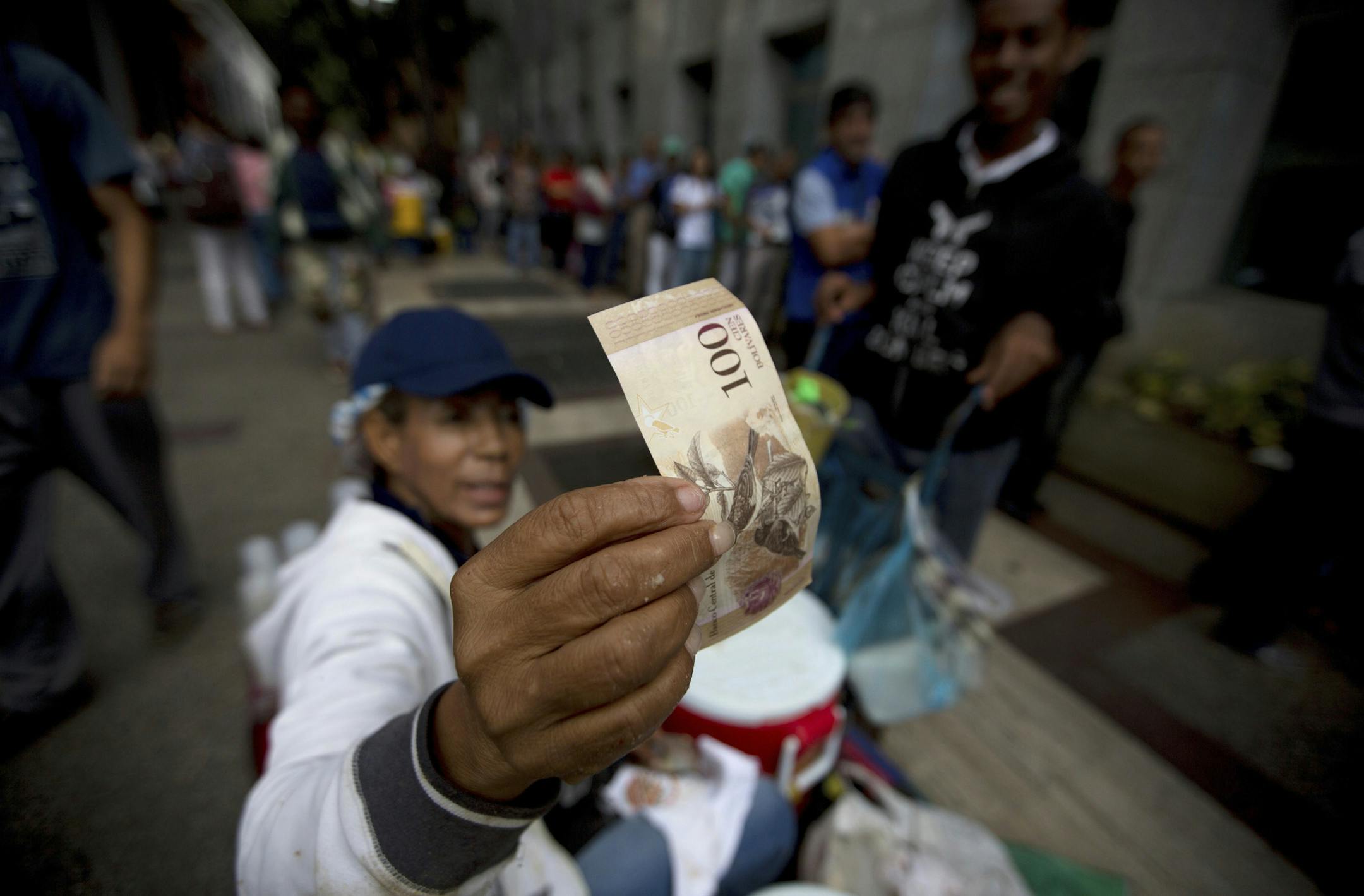 A street vendor inspects the authenticity of a 100-bolivar note as people stand in line outside a bank to deposit their bank 100-bolivar bank notes, in Caracas, Venezuela, Tuesday, Dec. 13, 2016. Venezuelans are taking a break from lining up to buy food and medicine to wait in long lines to deposit bank notes that have suddenly been declared worthless. President Nicolas Maduro made a surprise announcement this week that the 100-bolivar note, the country's largest-denominated bill and by far the