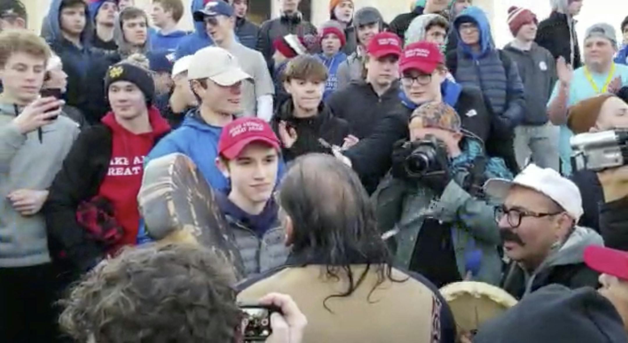 In this Friday, Jan. 18, 2019 image made from video provided by the Survival Media Agency, a teenager wearing a "Make America Great Again" hat, center left, stands in front of an elderly Native American singing and playing a drum in Washington. The Roman Catholic Diocese of Covington in Kentucky is looking into this and other videos that show youths, possibly from the diocese's all-male Covington Catholic High School, mocking Native Americans at a rally in Washington. (Survival Media Agency via