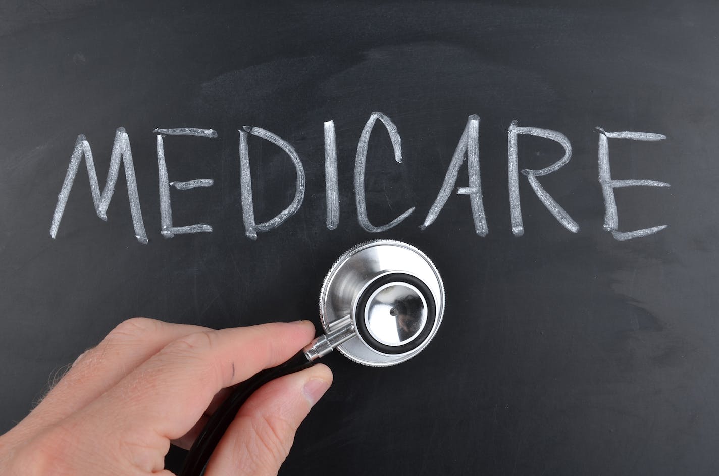 Medicare written on a blackboard in white chalk with a doctor using a stethoscope to check up.