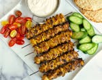 A platter with chicken shawarma skewers, tomatoes and cucumbers served with a side of dipping sauce and pitas.