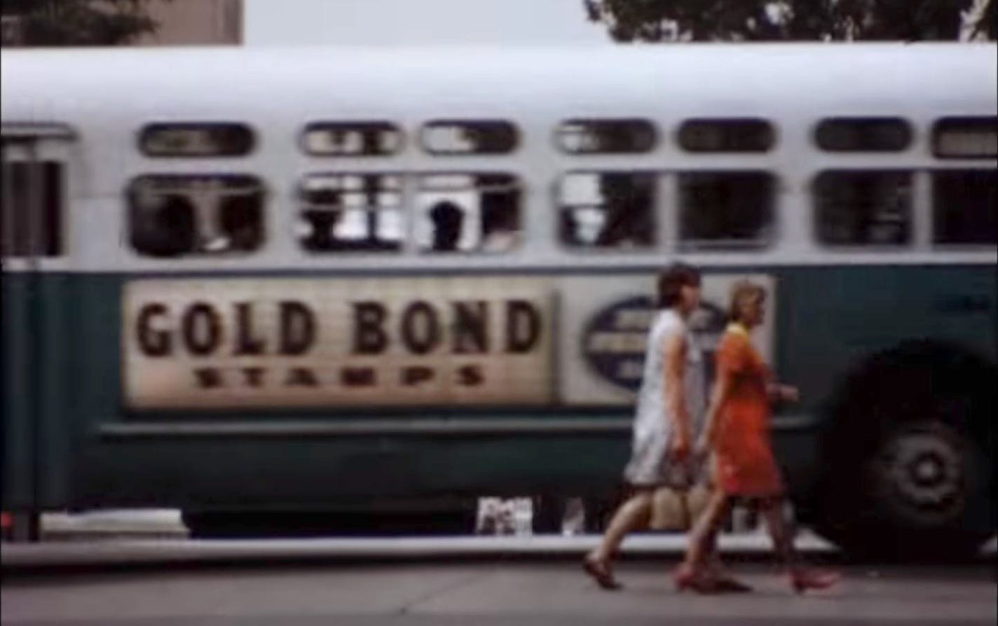 Photo credit:Augsburg University Archives
Still from lost Minneapolis promotional film. Two women walking as a bus emblazoned with Gold Bond advertising on the side passes by.