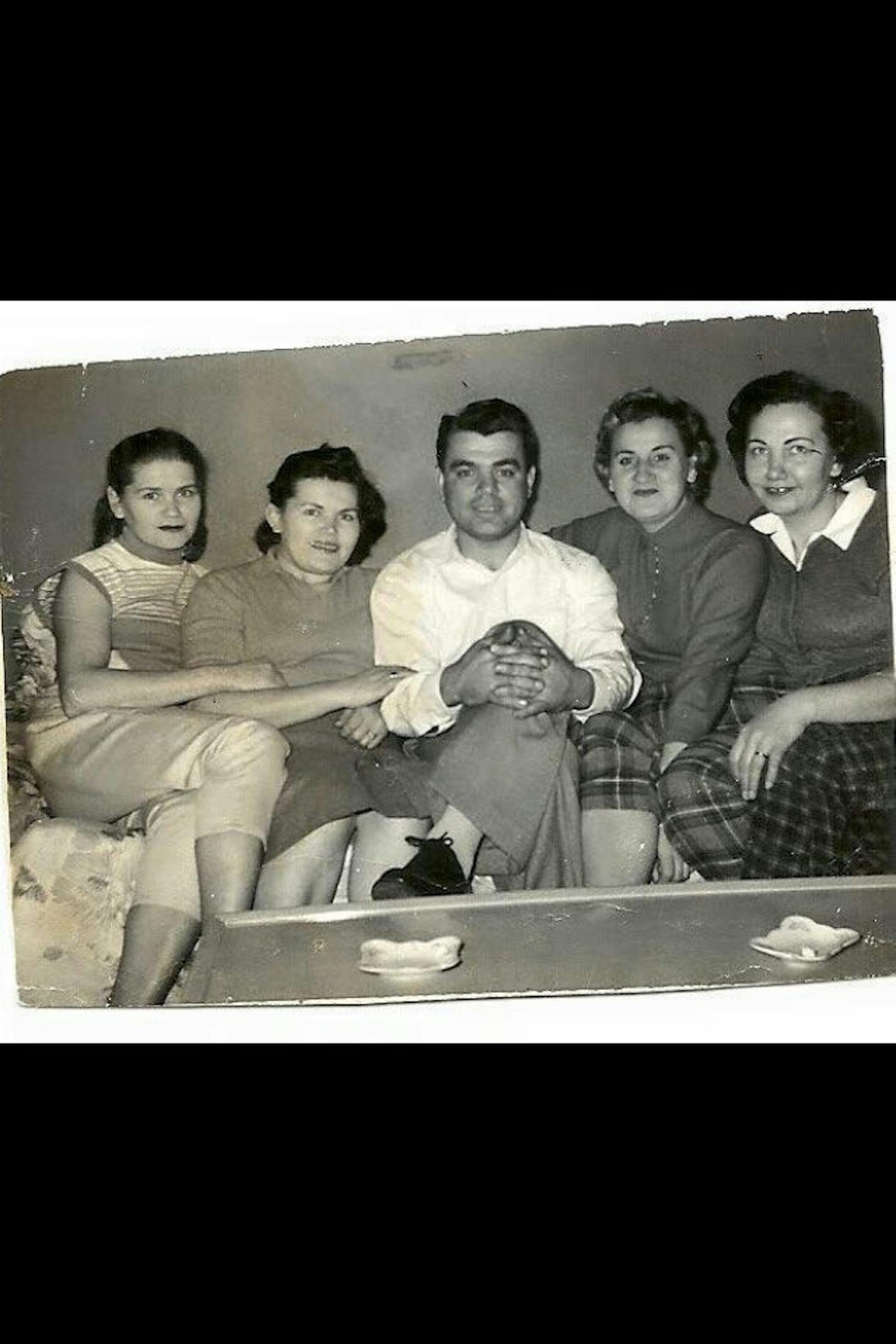 A black-and-white photo shows four women and a man sitting on the couch dressed in attire likely from the 1950s.