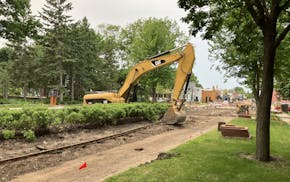 A yellow backhoe pulls long pieces of rail track from an unpaved roadway. Small shops are visible in the background.