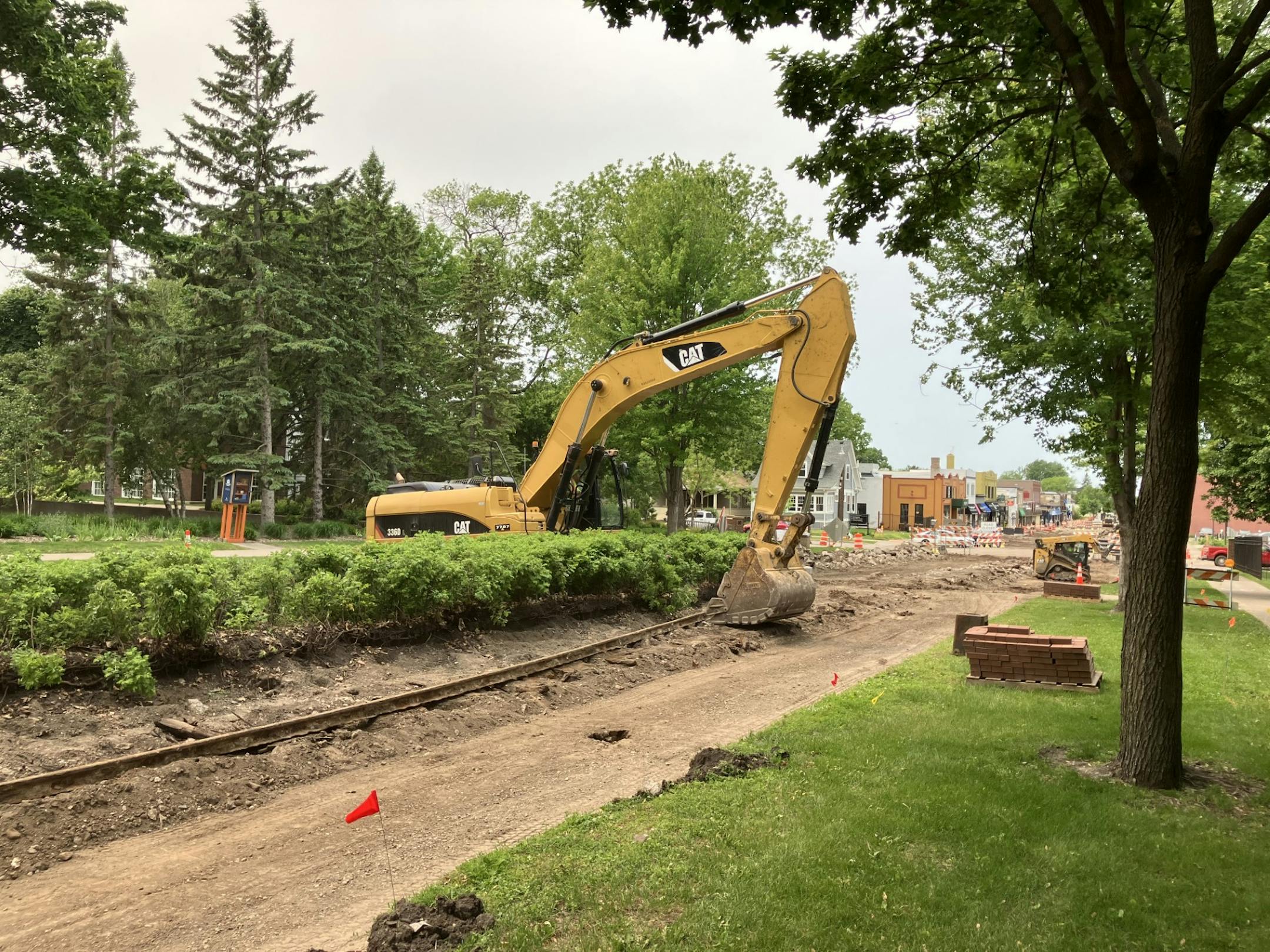 A yellow backhoe pulls long pieces of rail track from an unpaved roadway. Small shops are visible in the background.