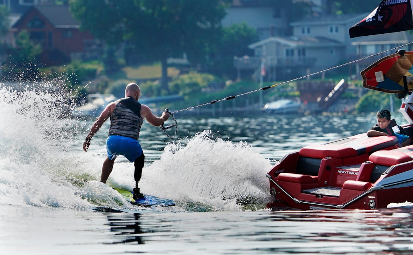 The 2021 Minnesota Wakesurf Championship, which runs through Saturday on Lake Minnetonka, kicked off Friday with two wake surfing group events. Joining together for a morning of wake surfing near Surfside Park were diasabled veterans from Wake for Warriors and a group for children from the metro's Urban Ventures joining Inner City Surf. Here, Marine Dustin Fleming, of San Antonio, TX, who lost a leg during combat to a bullet from a sniper, wake boarded on Lake Minnetonka while being watched by J.J. Brinson, 11, of Minneapolis with Urban Ventures who later waked boarded with Inner City Surf. ]