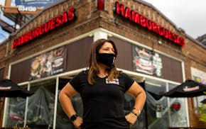 Claudia Gutierrez Mendez wearing black T-shirt and black face mask standing outside Hamburguesas El Gordo restaurant in Minneapolis with her hands on 