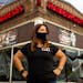 Claudia Gutierrez Mendez wearing black T-shirt and black face mask standing outside Hamburguesas El Gordo restaurant in Minneapolis with her hands on 