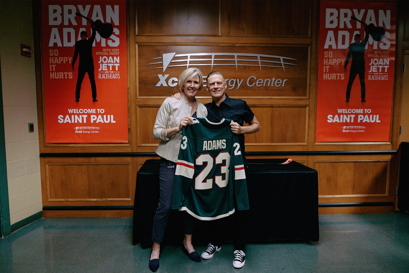 Xcel Energy Center's Kelly McGrath presents rock star Bryan Adams with a Minnesota Wild jersey on July 3, 2023 as they stand backstage in front of concert posters, posing for a photo.