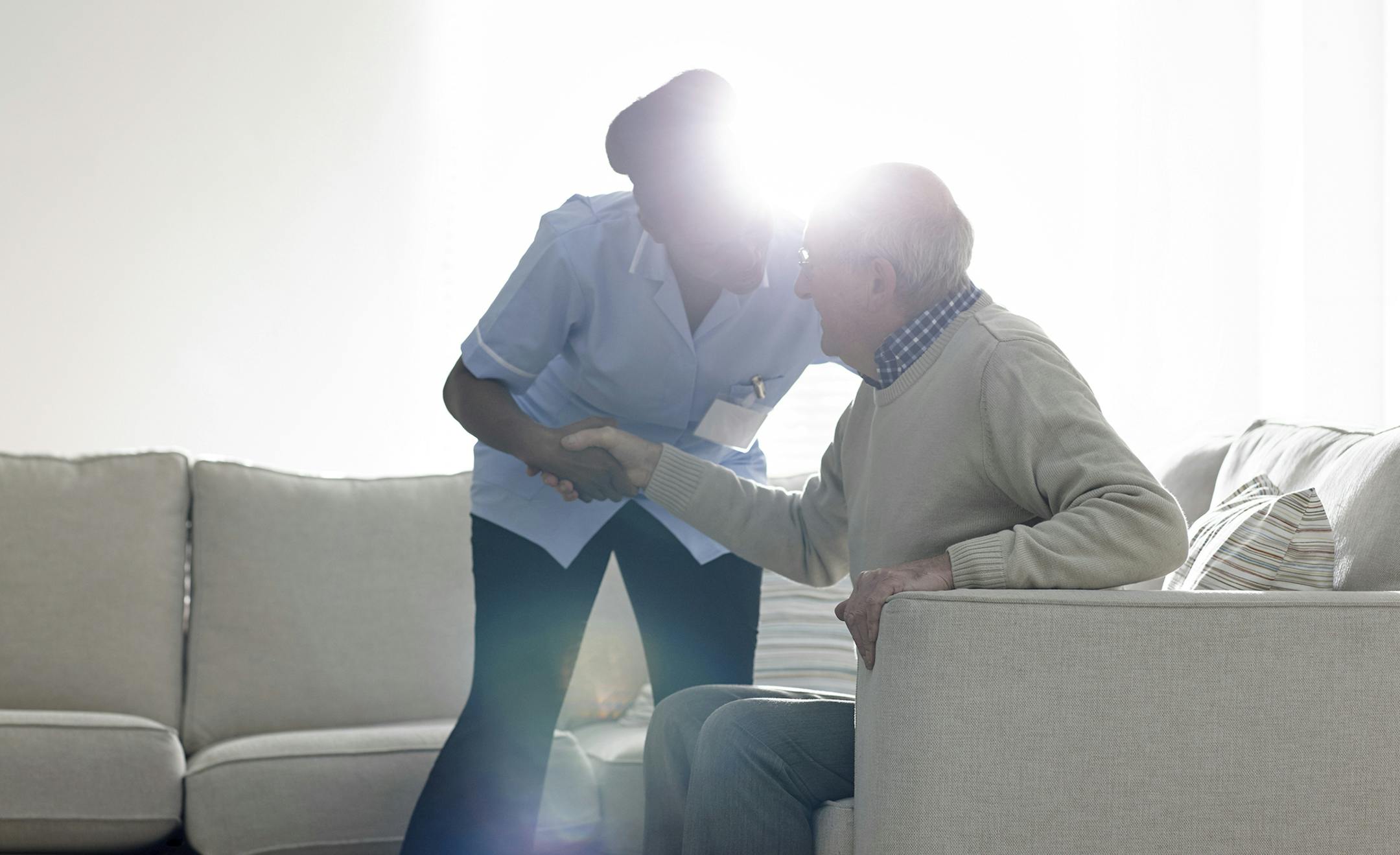 Female caregiver helping senior man get up from sofa at home