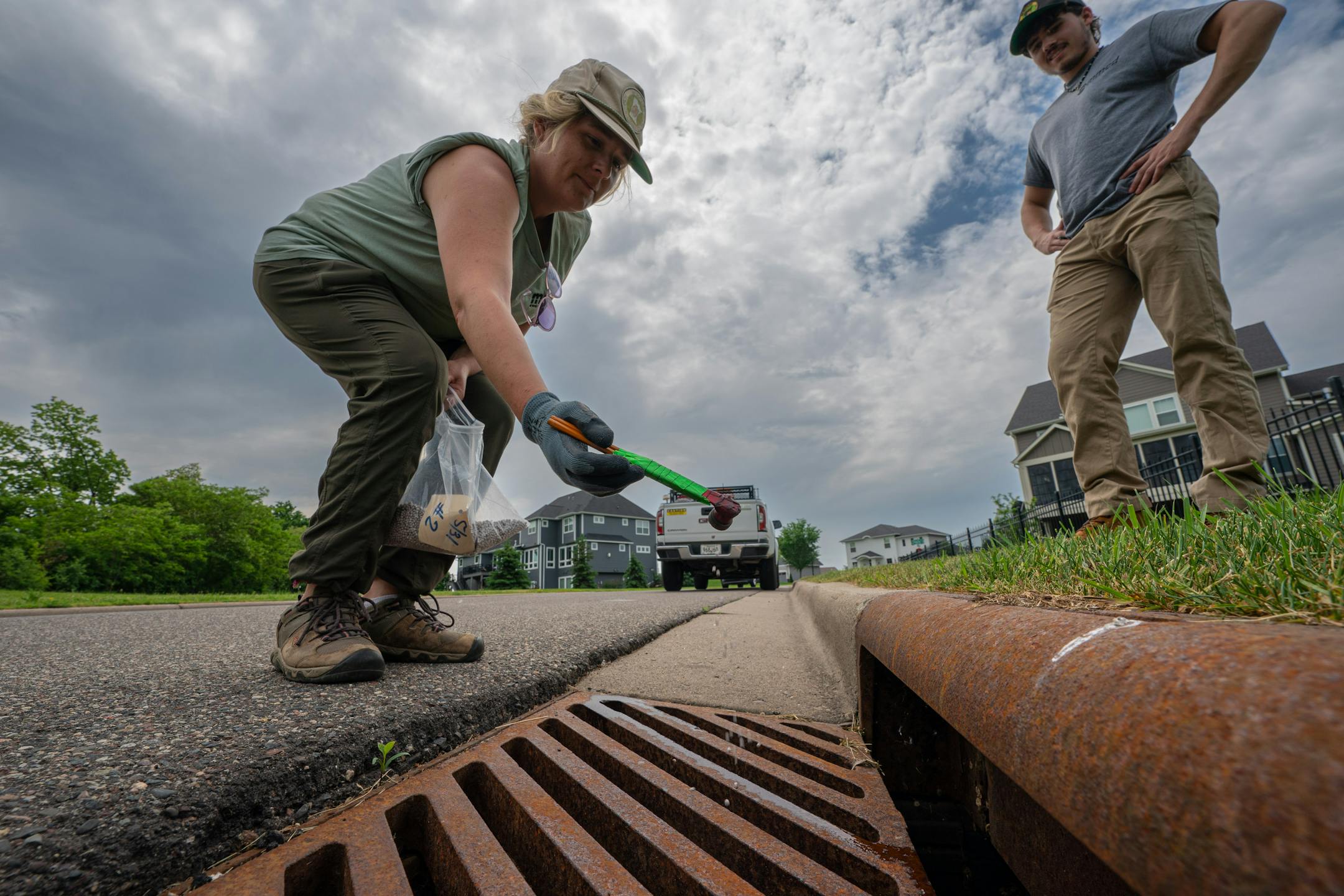 Field technician Gabriella Chacon pured a measured scoop of anti mosquito pellets into a storm drain near Pomerleau Lake, on the right is Ethan Nedina who just started with mosquito control.